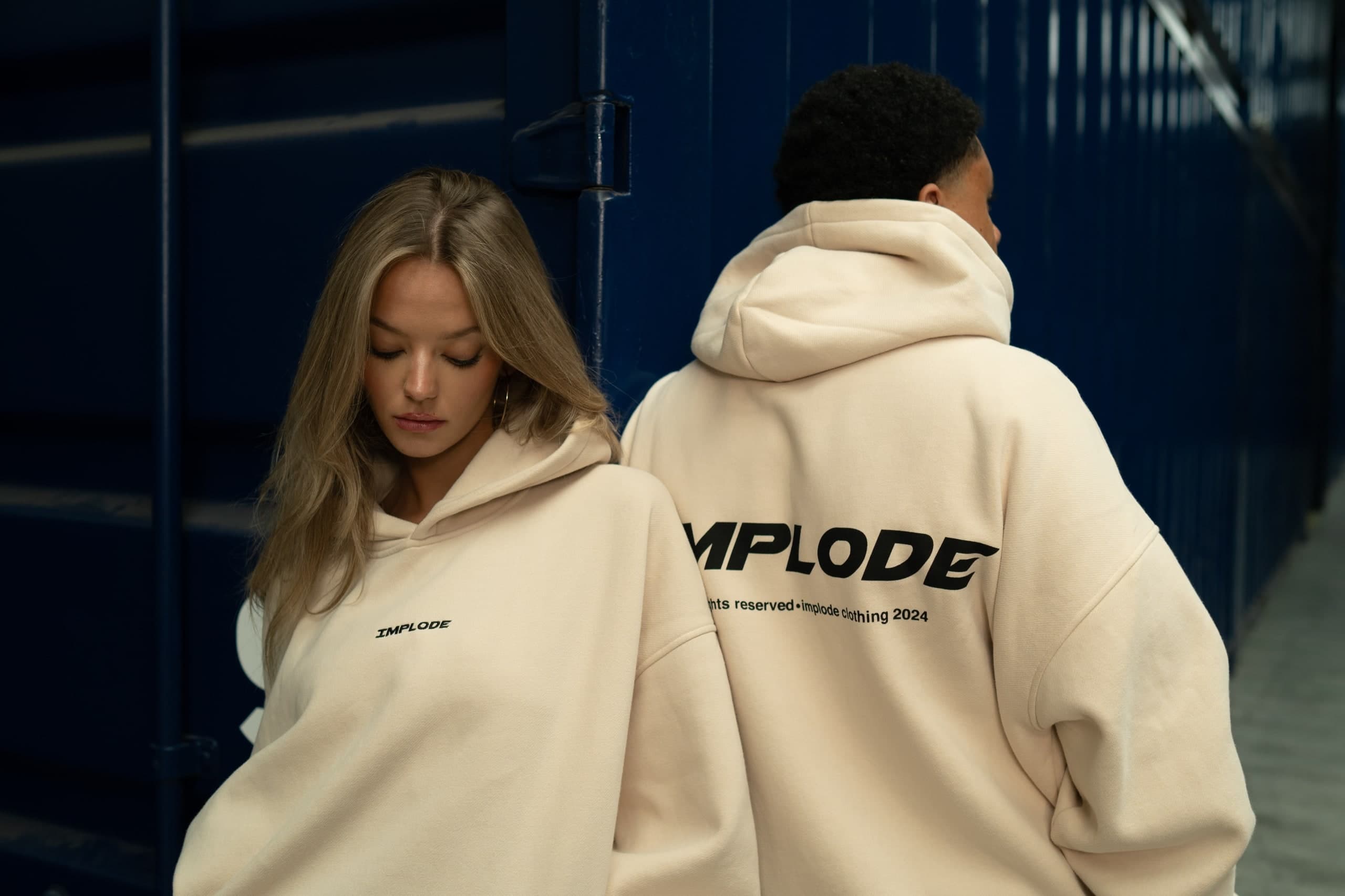 Zwei Personen in cremefarbenen Hoodies vor einer dunkelblauen Wand